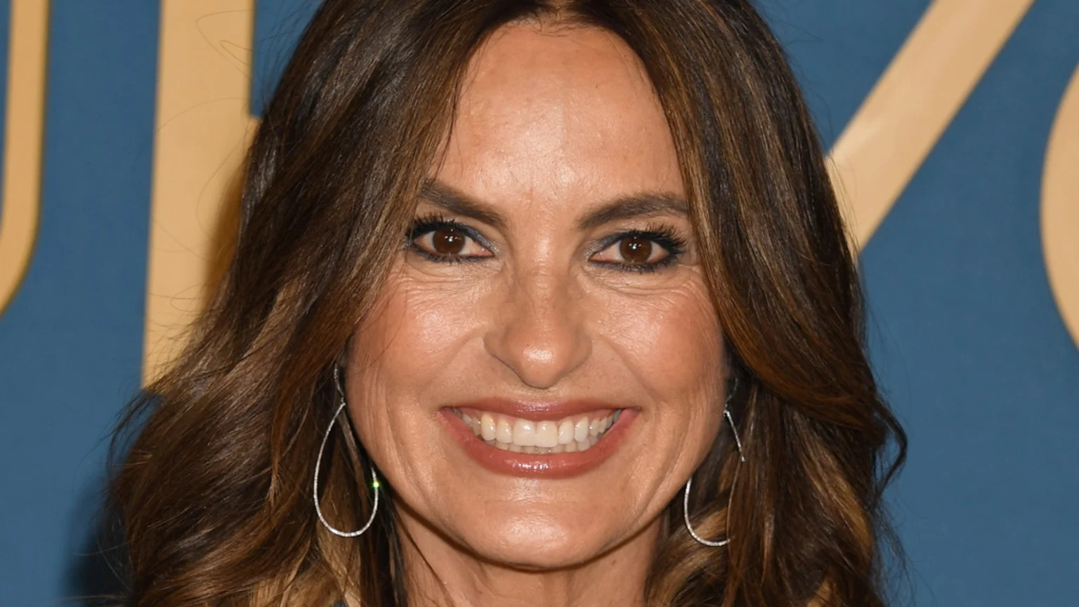 A close-up portrait of Mariska Hargitay smiling widely, showing her teeth, with long brown hair and teardrop earrings, against a blurred blue and gold background.