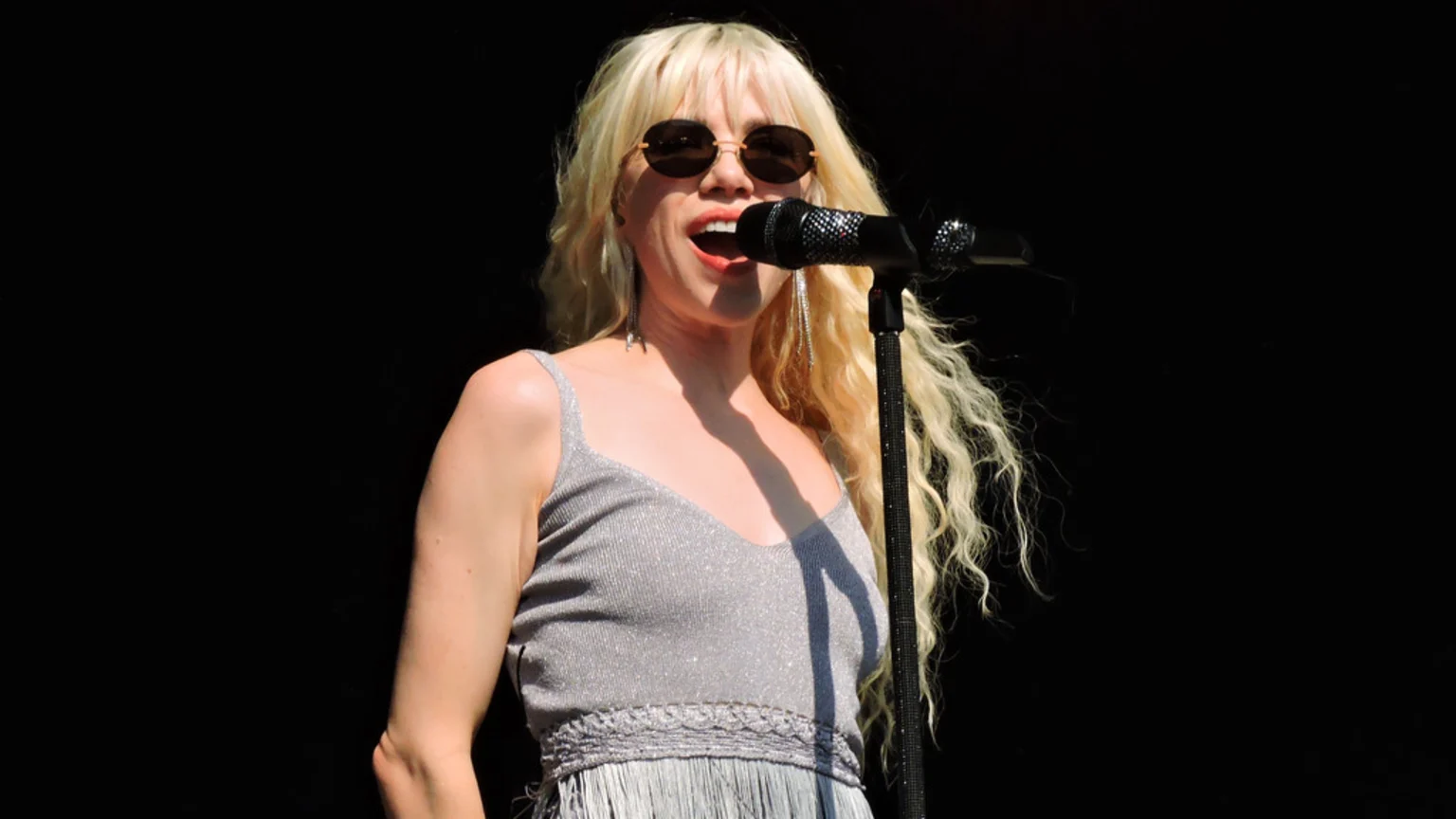 A woman with long, wavy blonde hair and sunglasses sings into a microphone, wearing a shimmering silver V-neck top with thin straps and fringe, against a dark background.