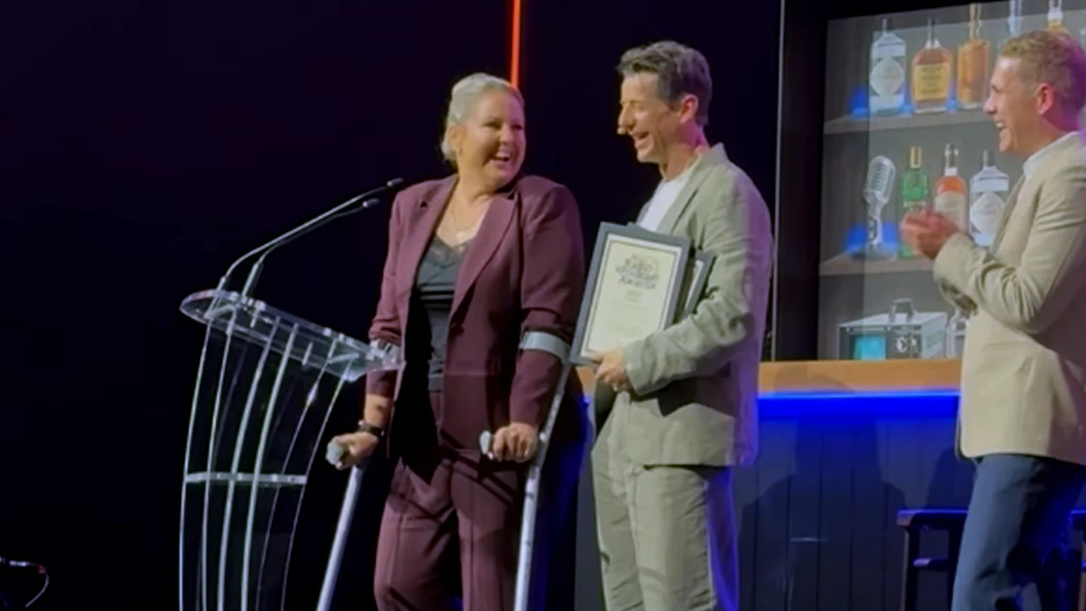 A smiling woman on crutches in a maroon suit stands next to a man holding awards, who smiles back at her on stage, as a third man claps in the background.