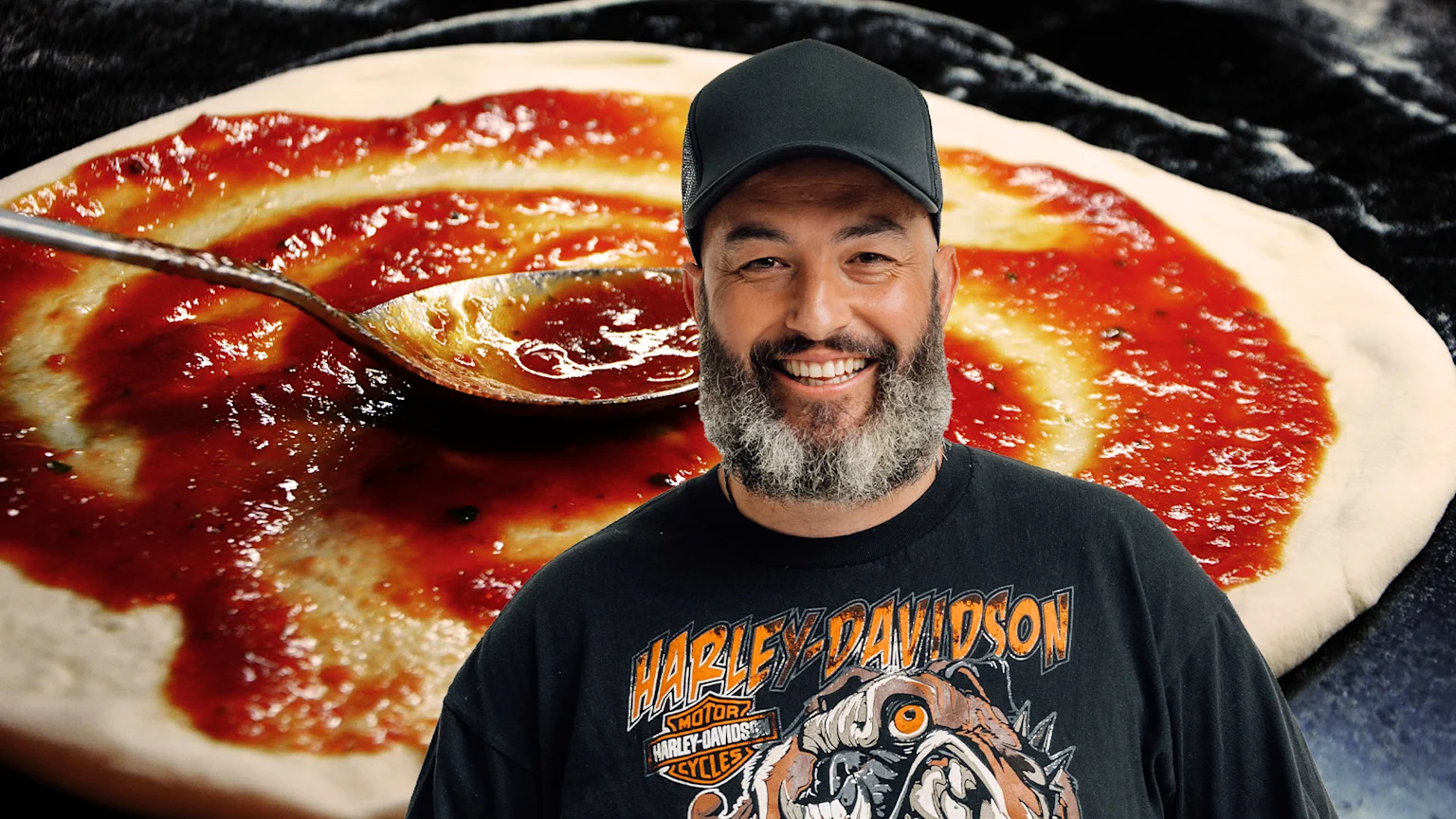 A smiling man in a black cap and Harley-Davidson shirt stands in front of a pizza dough being spread with red sauce by a spoon.
