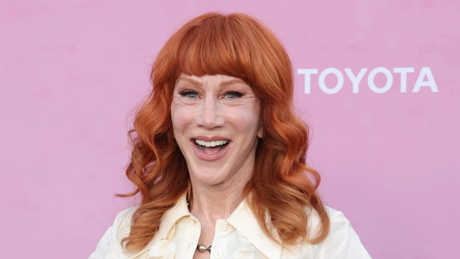 Kathy Griffin, a woman with red hair and bangs, smiles widely against a pink background with a white Toyota logo visible.