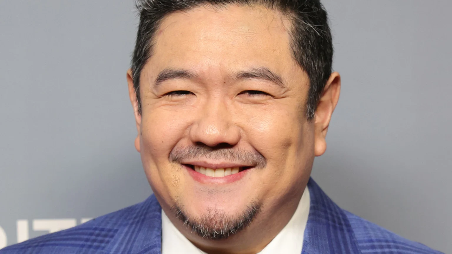 A smiling East Asian man with dark hair, a mustache and goatee, wearing a blue plaid suit jacket over a white shirt.
