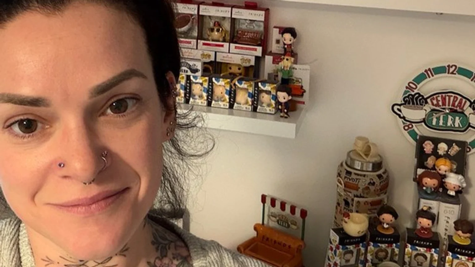 A smiling woman with multiple piercings and tattoos poses in front of a shelf filled with "Friends" themed merchandise, including figurines, a Central Perk clock, and the iconic orange couch.