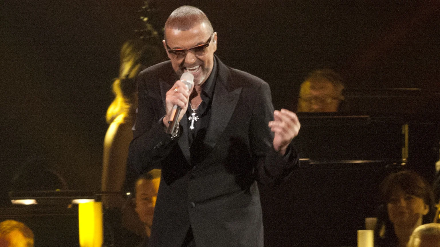 George Michael, wearing sunglasses and a black suit, smiles and sings into a microphone on stage, with blurred figures visible in the dark background.