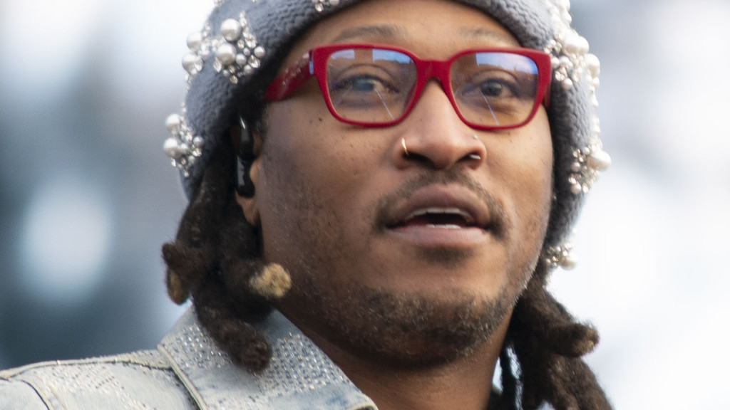 A close-up of Rapper Future shows him wearing red glasses, a grey beanie adorned with pearls and rhinestones, a nose ring, and dreadlocks, looking slightly upwards with his mouth slightly open.