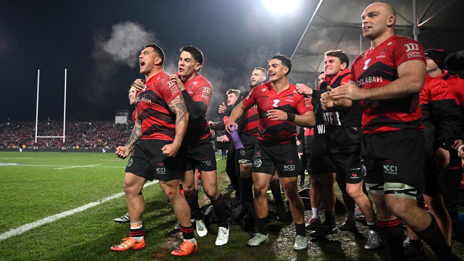 A group of male rugby players in red and black uniforms cheer and walk off a rugby field at night, with one player shouting and visible breath in the cold air, and stadium lights and goalposts in the background.