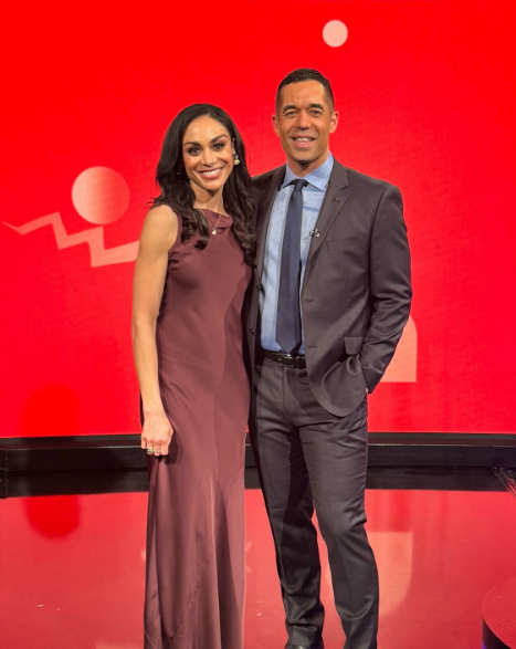 A smiling woman in a long maroon dress and a man in a dark suit with a microphone on his tie stand side-by-side in front of a red background with white abstract shapes.