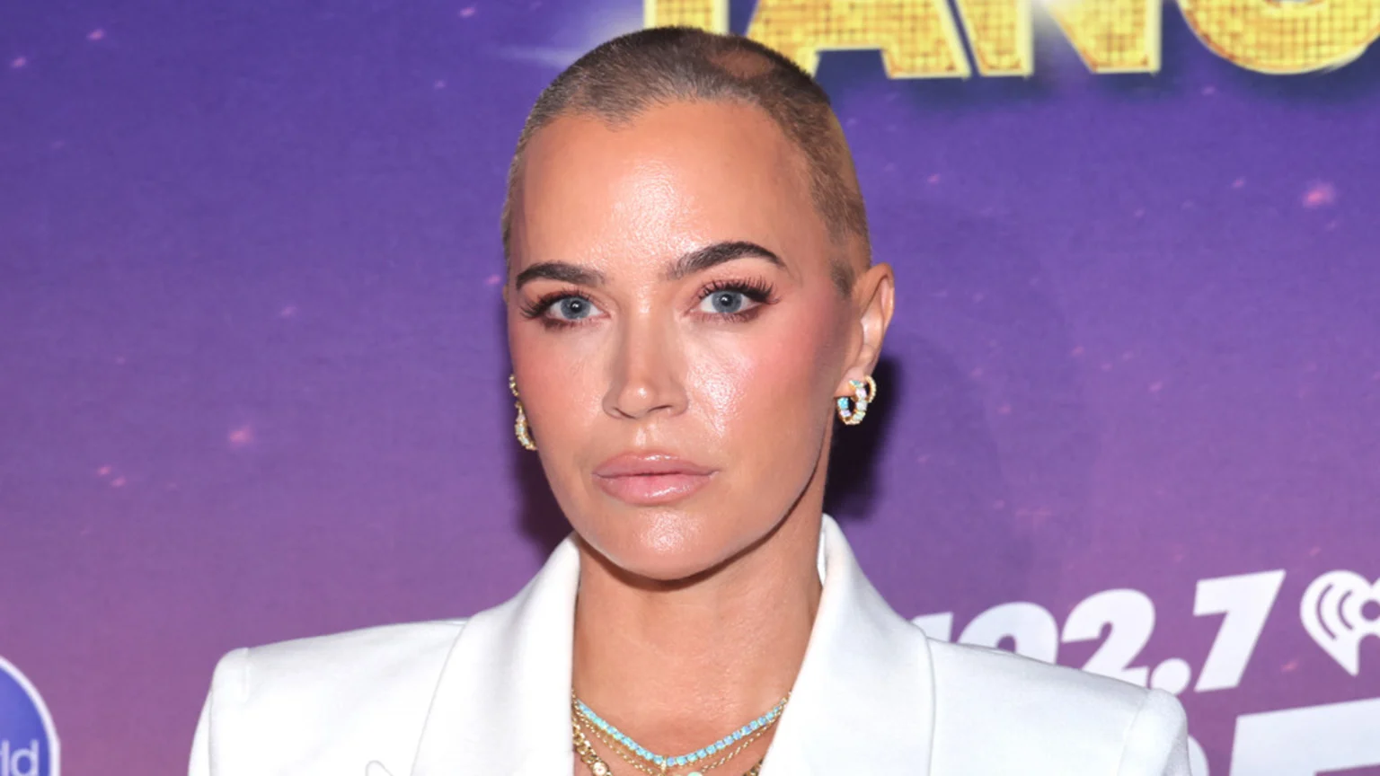 A woman with a very short buzz cut, full makeup including blue eyes and prominent lashes, and a white blazer looks directly forward against a blurry purple background.