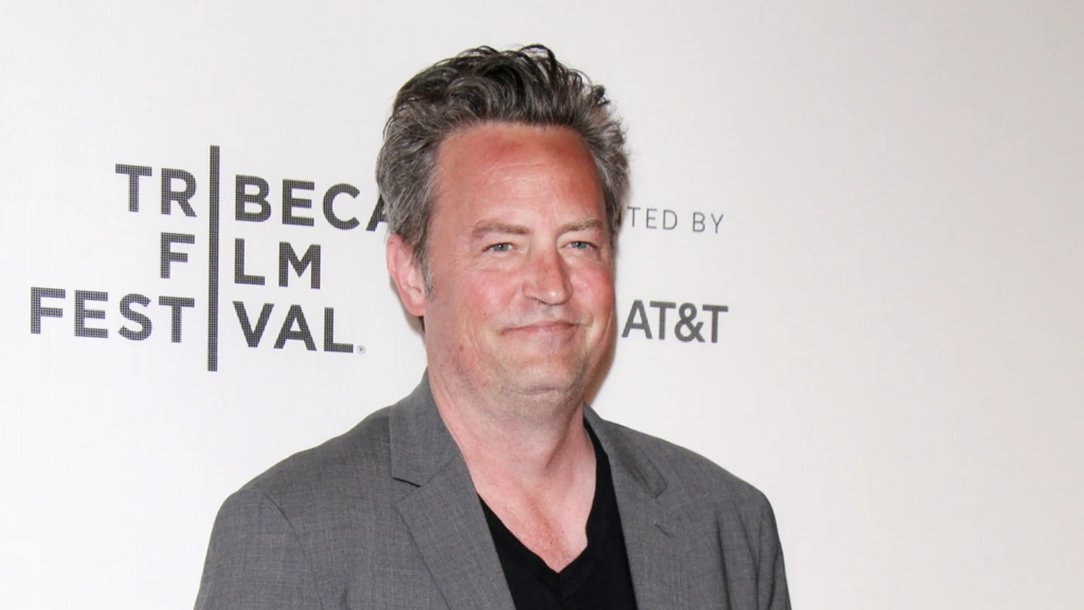 Matthew Perry smiles slightly at the camera, wearing a grey blazer and black V-neck shirt, in front of a Tribeca Film Festival backdrop.