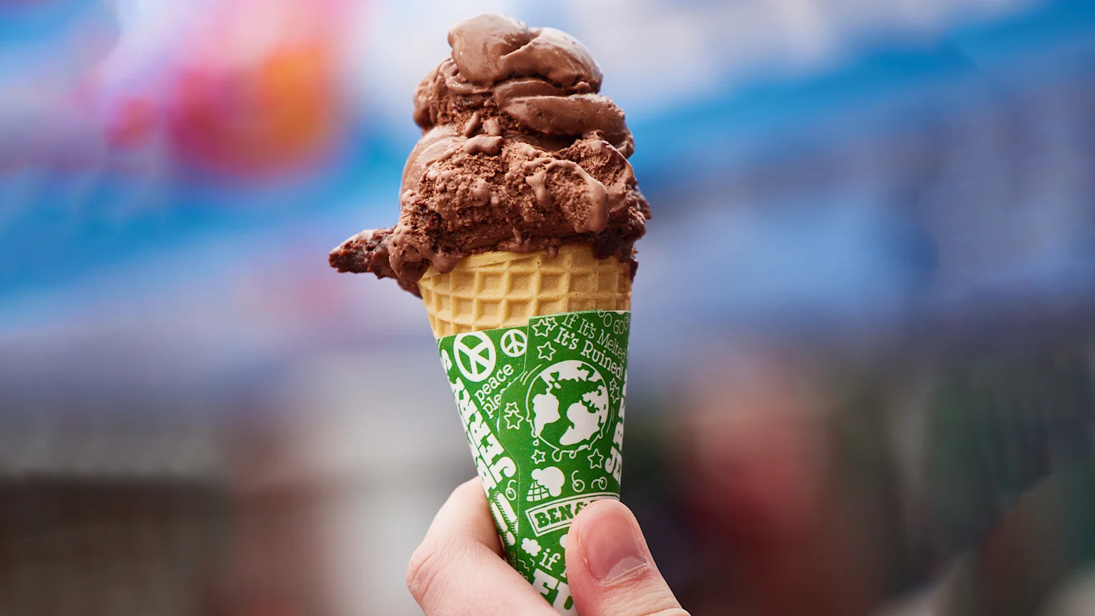 A hand holds a chocolate ice cream cone with a green Ben & Jerry's wrapper, featuring a peace sign and an earth globe, against a blurred, colorful background.