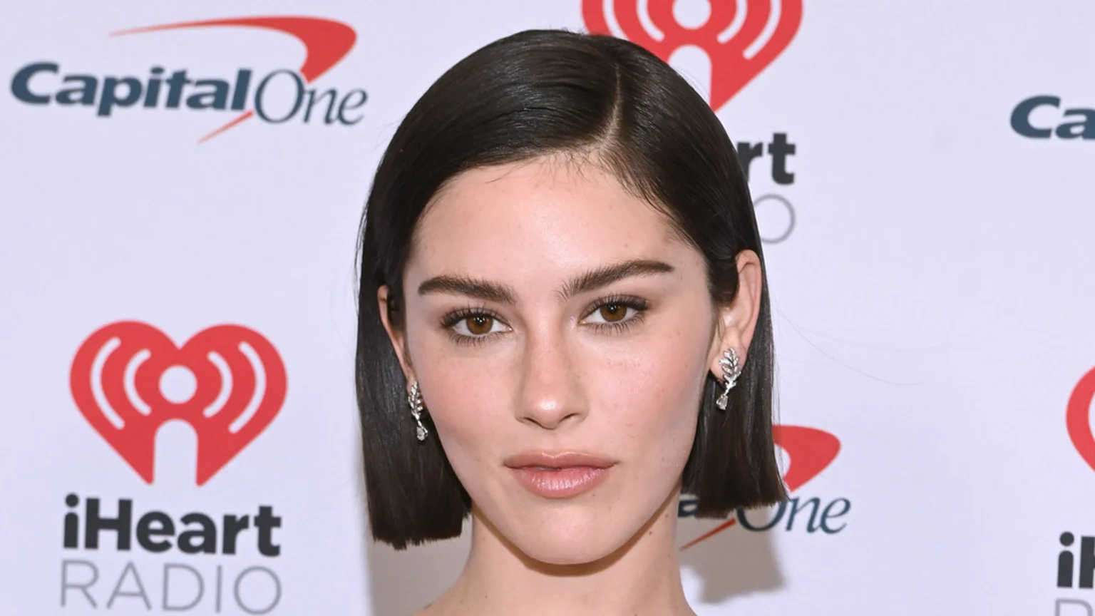 Gracie Abrams, a young woman with dark brown hair, brown eyes, and bold eyebrows, looks directly at the camera, wearing leaf-shaped dangle earrings against a background of white logos.
