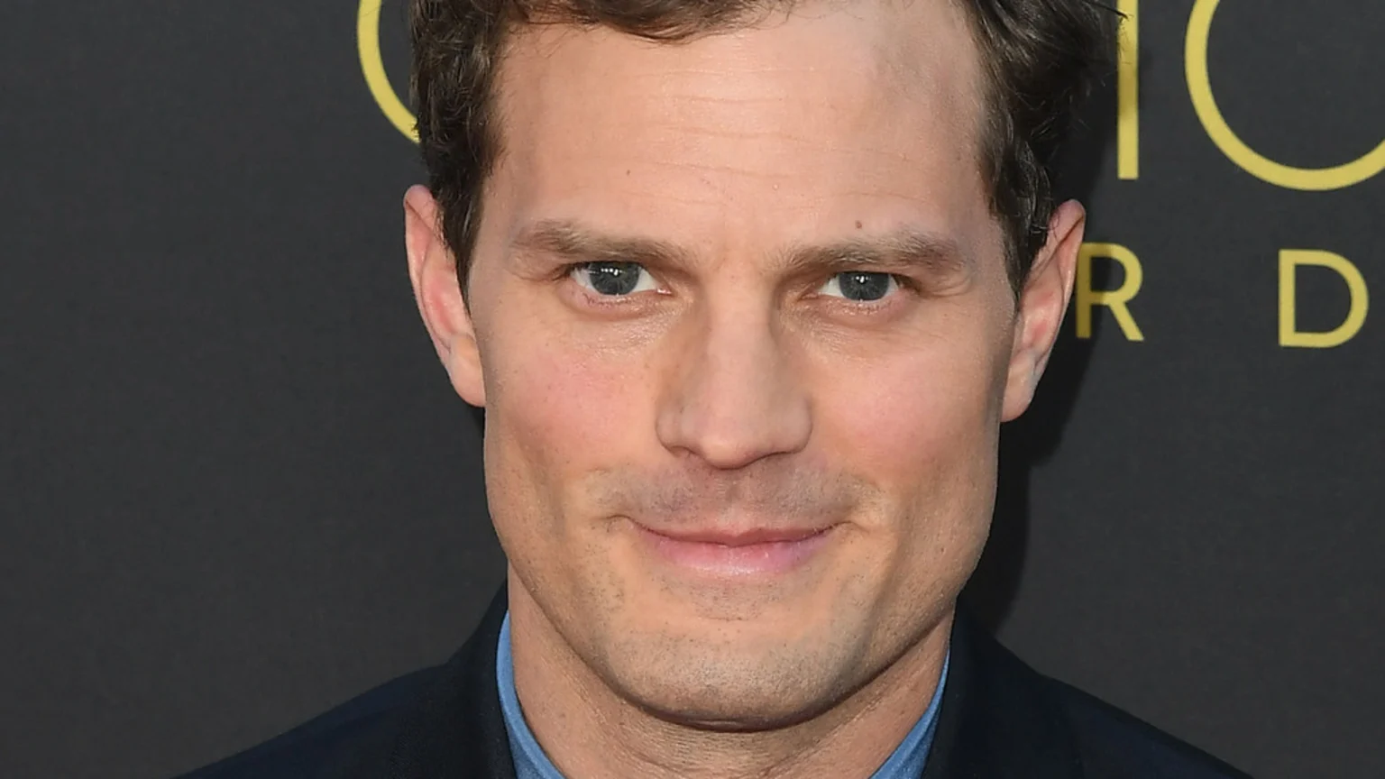 A close-up portrait of a light-skinned man with light brown hair and blue eyes, wearing a dark suit jacket over a blue shirt, looking directly at the camera with a subtle smile.