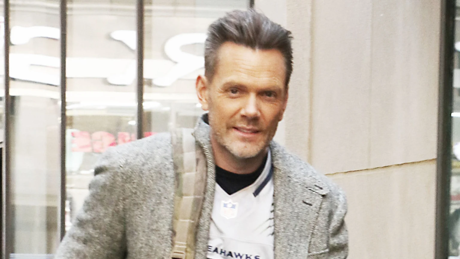 Joel McHale, with short hair and a light beard, smiles at the camera, wearing a grey coat over a white Seattle Seahawks NFL jersey, with a blurred background.