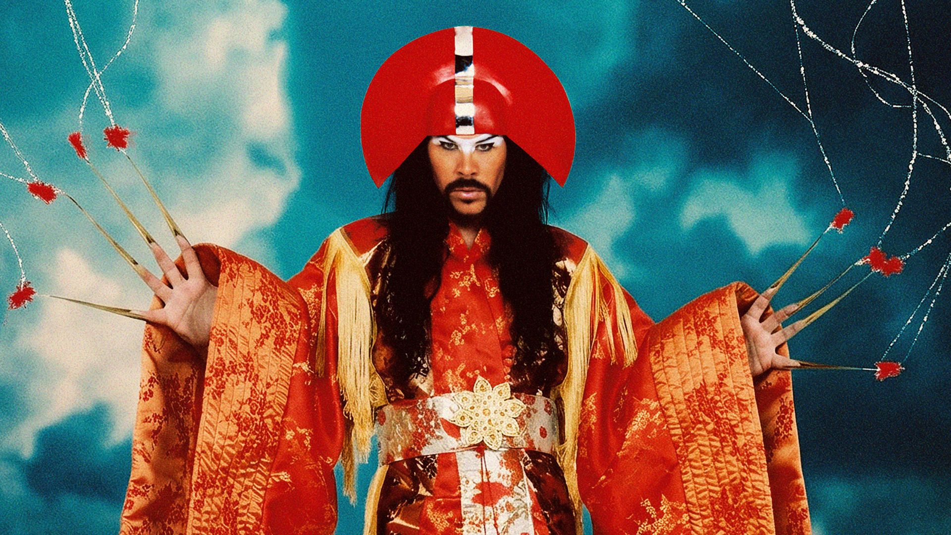 A person in an elaborate red and gold robe with a red headdress, dark hair, beard, and dramatic makeup stands against a blue sky, displaying long golden finger claws connected to sparkling threads with red flowers.