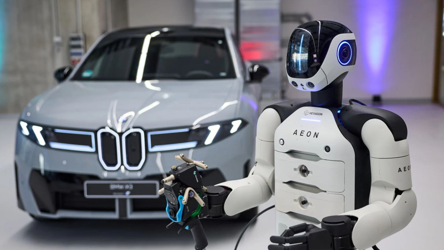 A white and black Hexagon AEON humanoid robot holds a sensor device, standing in front of a light gray BMW iX3 car with its headlights illuminated.