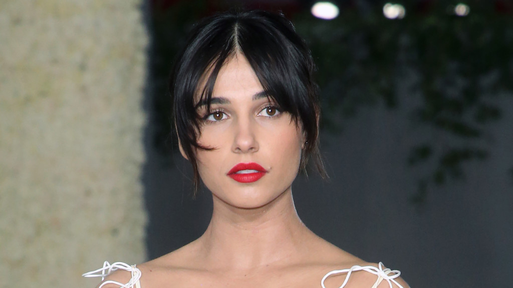 A headshot of Naomi Scott, with dark hair and wispy bangs, wearing red lipstick and a light-colored top with delicate white string details.
