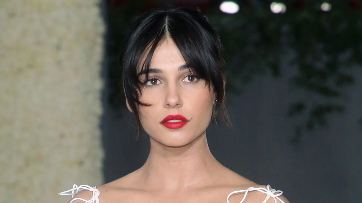 A headshot of Naomi Scott, with dark hair and wispy bangs, wearing red lipstick and a light-colored top with delicate white string details.