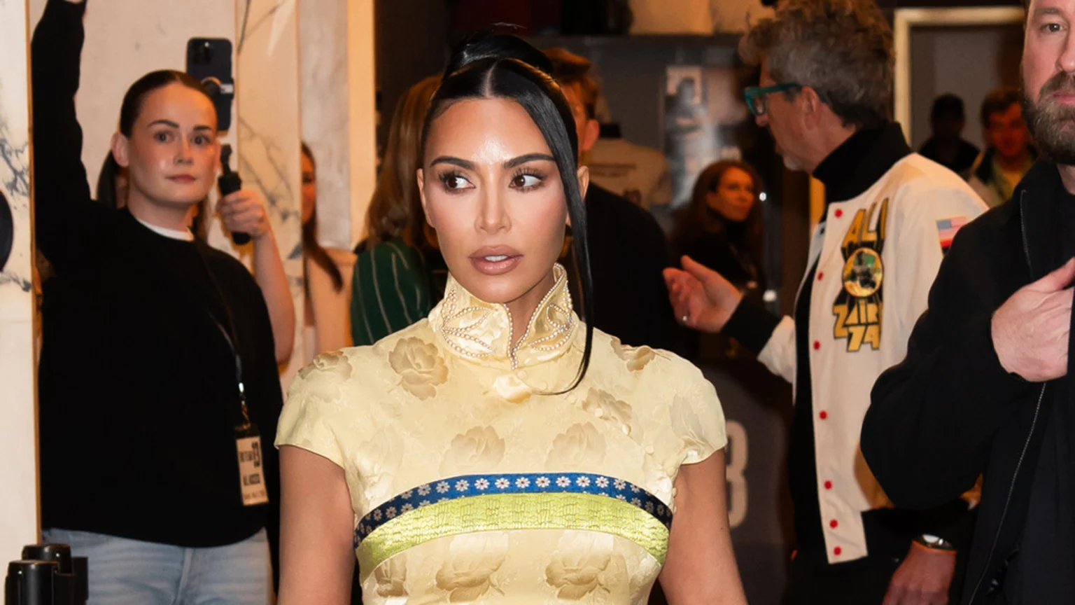 Kim Kardashian in a yellow floral dress with a high pearl-decorated collar and a blue and green band, her hair in a sleek high ponytail, looking right, with people in the background.