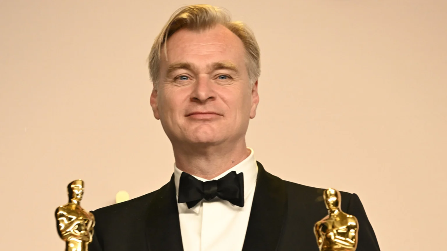 Christopher Nolan, a man with light hair wearing a tuxedo and bow tie, smiles while holding two golden Oscar statues against a plain background.