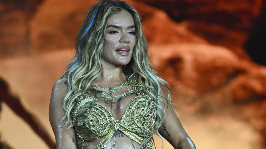Close-up of Karol G with long blonde hair and blue streaks, wearing a shimmering golden top, looking to her right with her mouth slightly open, on a stage with warm lighting.