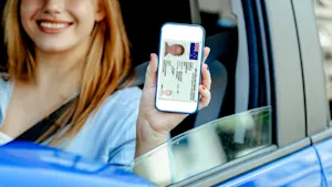 Blonde woman sitting in a blue car with her hand out the window revealing a New Zealand Driver's Licence on her phone, digitally.