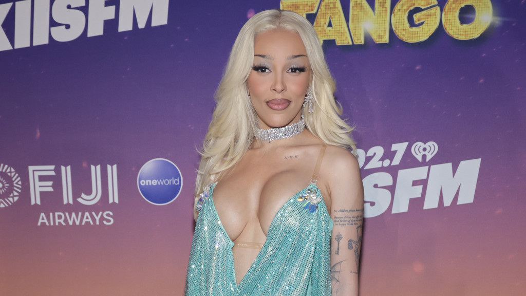 Doja Cat poses for a photo, wearing a shimmering turquoise dress with a deep neckline, a diamond choker, and visible tattoos, against a purple background with various logos.