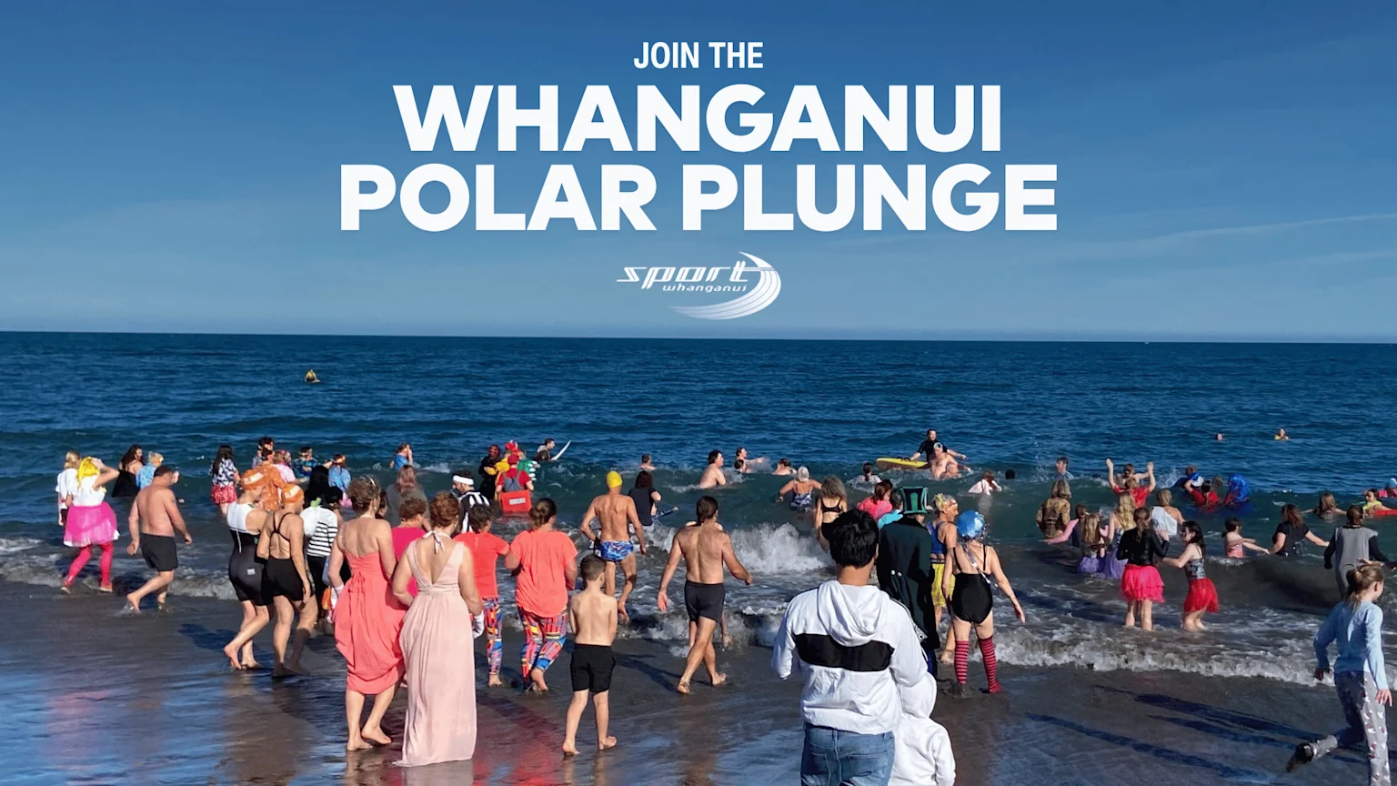 A crowd of people in swimsuits and costumes wades into the blue ocean water on a sunny day for the Whanganui Polar Plunge event.