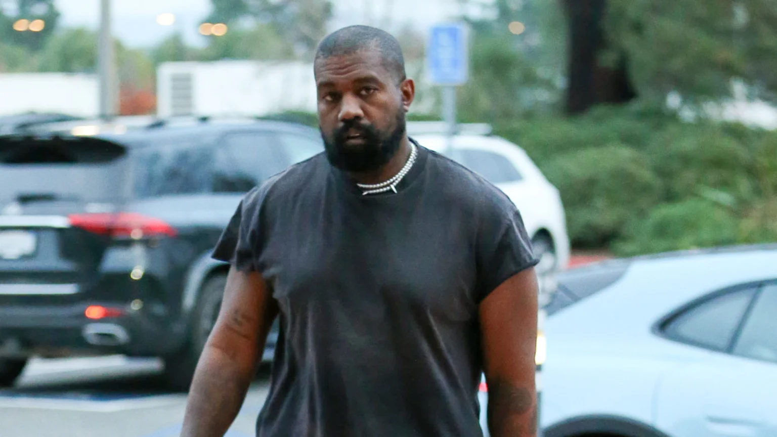 Kanye West with a beard and short hair wears a dark t-shirt and two silver chain necklaces, looking directly ahead, with blurred cars and greenery in the background.