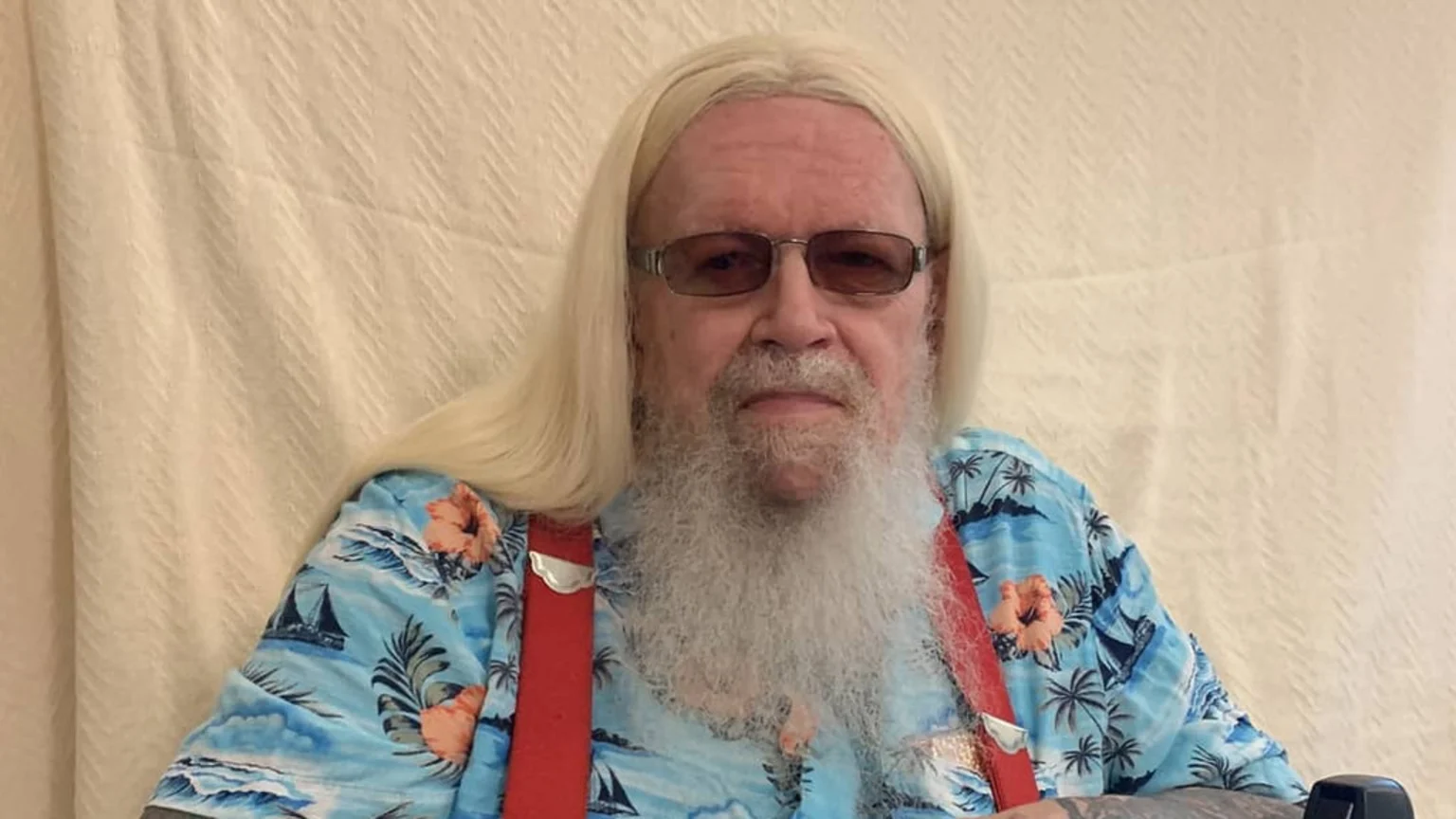 A man with long blonde hair, a long white beard, and sunglasses wears a blue Hawaiian shirt with red suspenders against a light background.