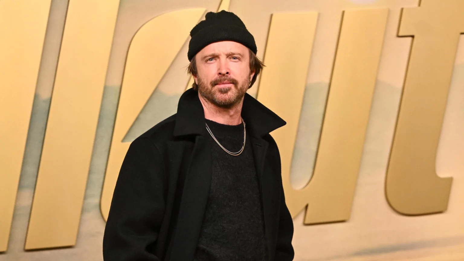 A man with a beard and black beanie smiles subtly, wearing a black coat, a black sweater, and two silver chain necklaces, standing in front of a blurred background with large, light-colored letters.