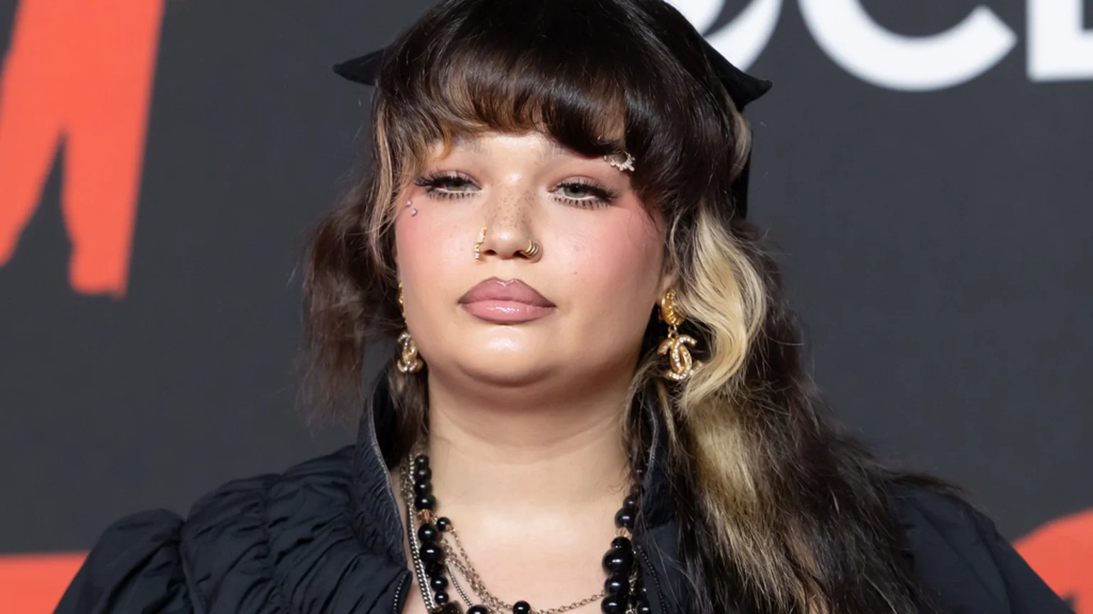 Close-up of Lola Young with dark hair and a blonde streak, wearing a black top, layered necklaces, and Chanel logo earrings, looking directly at the camera with noticeable facial piercings and makeup.
