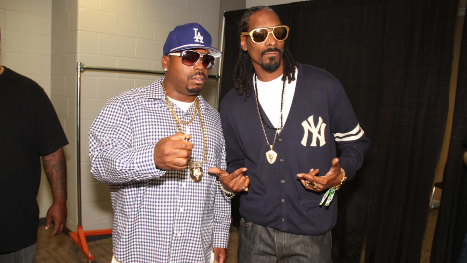 Daz Dillinger in a blue LA cap, sunglasses, and checkered shirt, holding a lighter, stands next to Snoop Dogg in gold sunglasses and a navy Yankees cardigan.