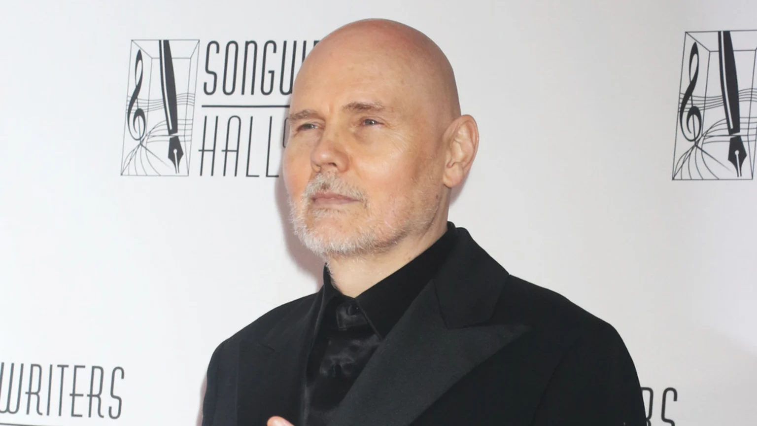Billy Corgan, bald with a gray beard, looks thoughtfully to the side while wearing a black suit jacket, with a "SONGWRITERS HALL" logo visible in the background.