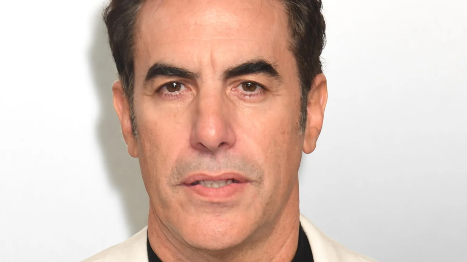 Close-up headshot of Sacha Baron Cohen, a man with dark hair, prominent dark eyebrows, and brown eyes, looking directly at the camera with a neutral expression against a plain white background.