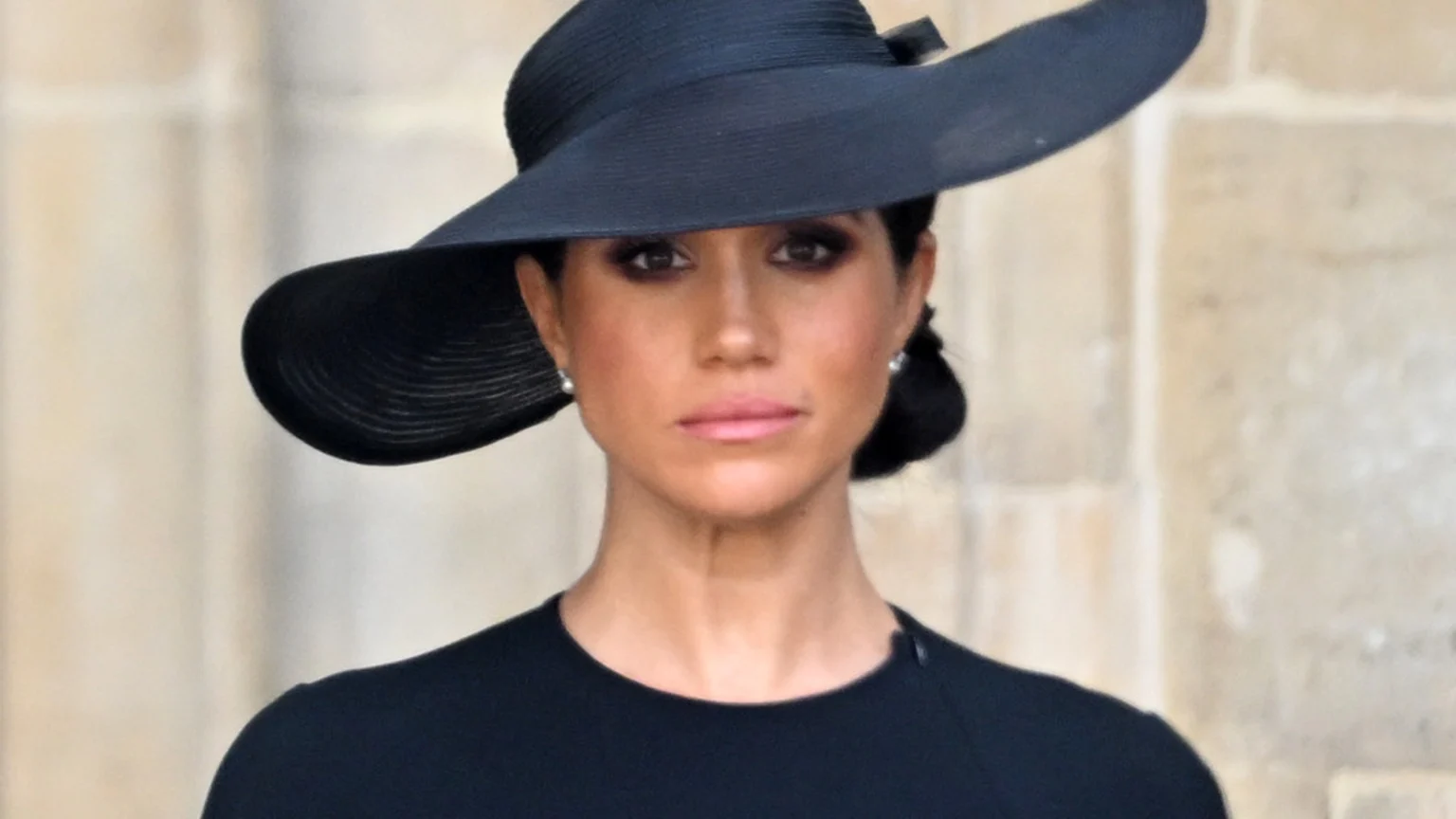 Meghan Markle wears a black wide-brimmed hat and black top, looking directly ahead with a somber expression, with pearl earrings visible.