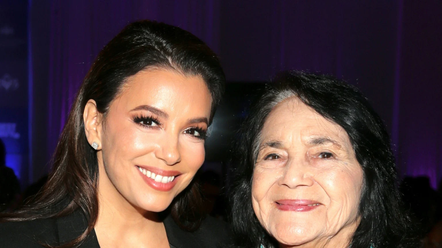 Eva Longoria, smiling in a black top, poses next to Dolores Huerta, an older woman with dark hair and a warm smile, against a blurry purple background.