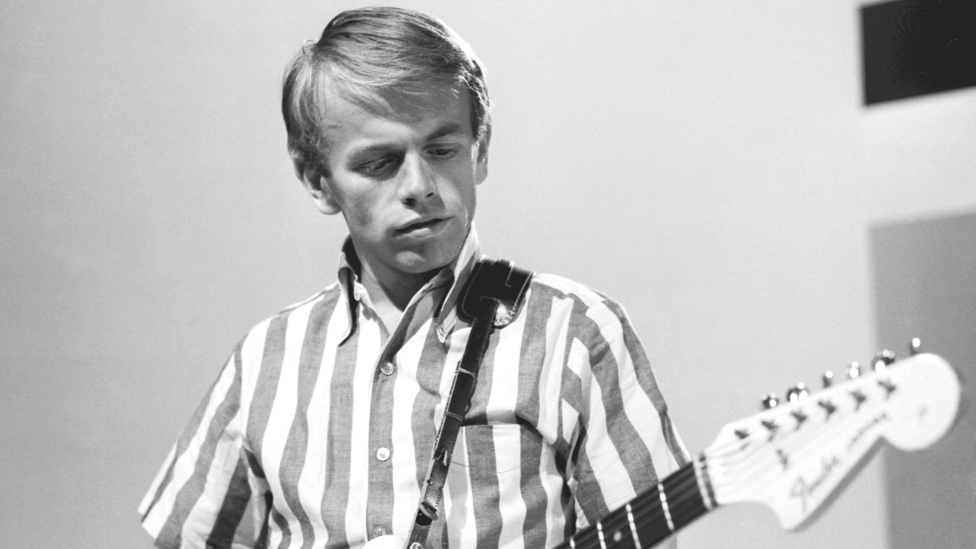 A black and white photo shows a man with short, light hair, wearing a striped short-sleeved shirt and a guitar strap, looking down while holding a Fender guitar.