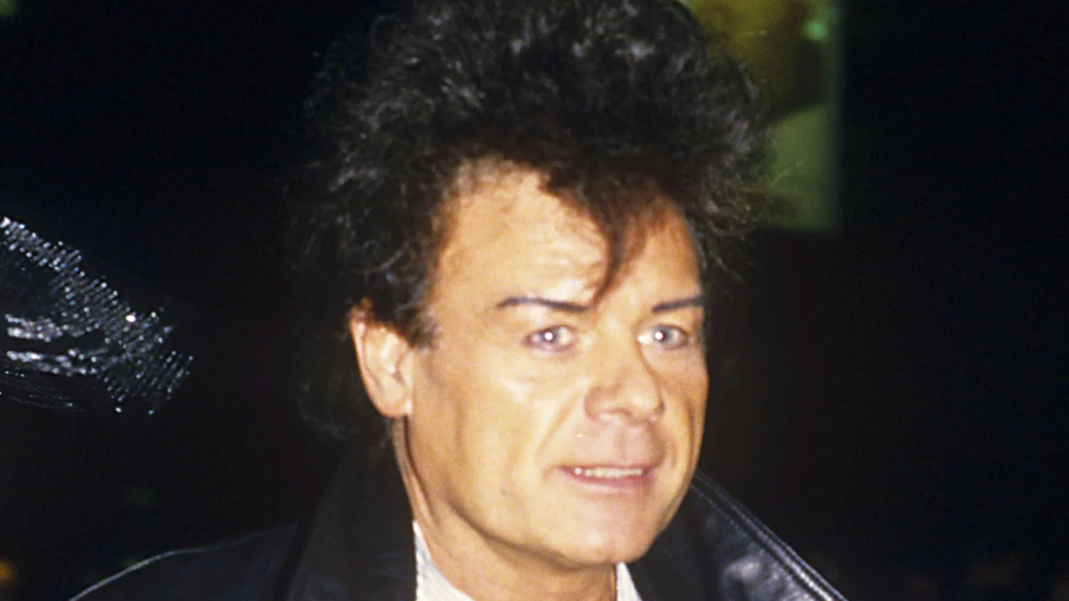 Close-up of Gary Glitter smiling slightly, featuring his dark, spiky hair, light eyes, and a black leather jacket, with a shimmering detail on the left.