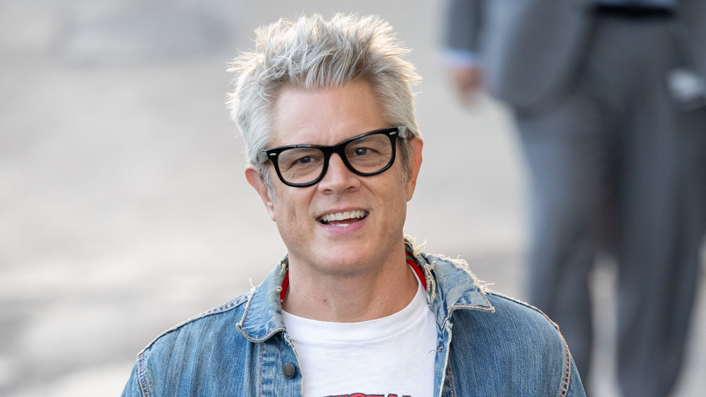 Johnny Knoxville, a man with spiky grey hair and black glasses, smiles directly at the camera, wearing a denim jacket over a white t-shirt.