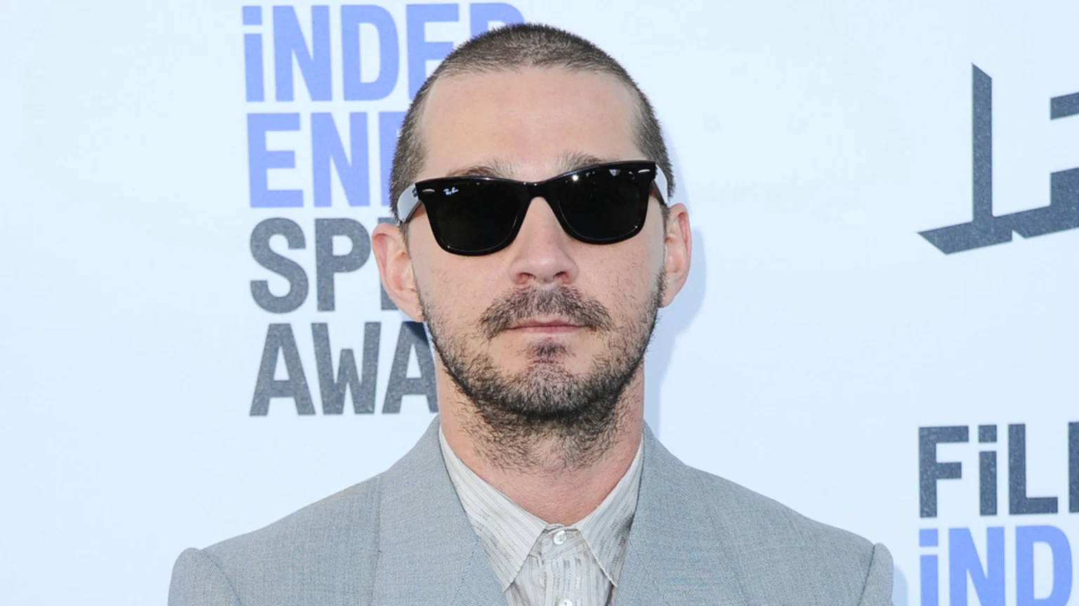 Shia LaBeouf with a buzz cut, beard, and black sunglasses, wearing a light grey suit, stands against a white banner.