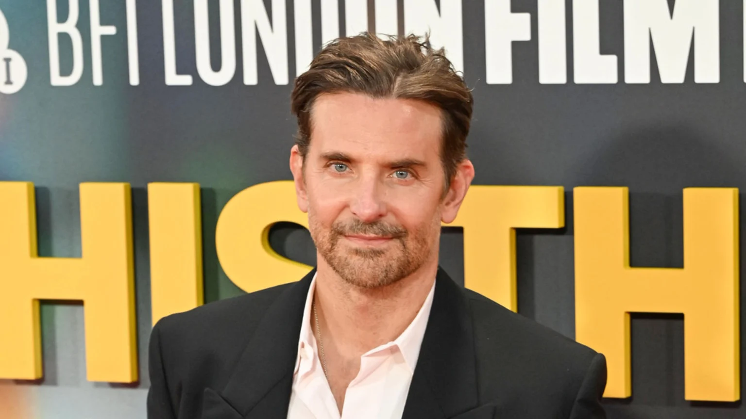 Bradley Cooper, with a beard and blue eyes, looks directly at the camera, wearing a dark suit and light shirt, in front of a blurred background with event signage.