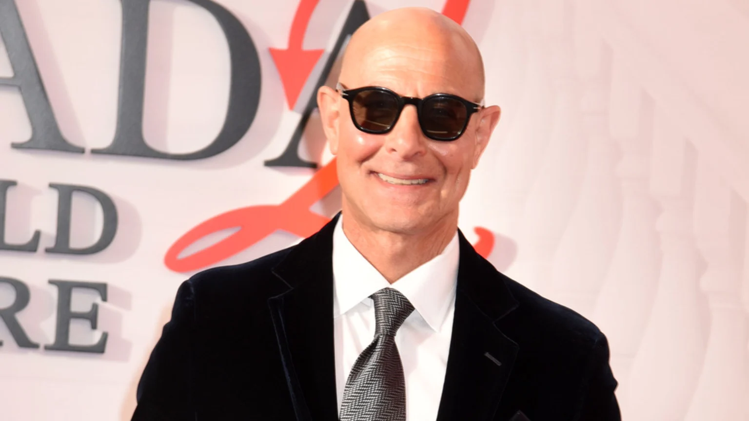 Bald Stanley Tucci smiles at the camera, wearing dark sunglasses, a dark velvet jacket, white shirt, and patterned tie.