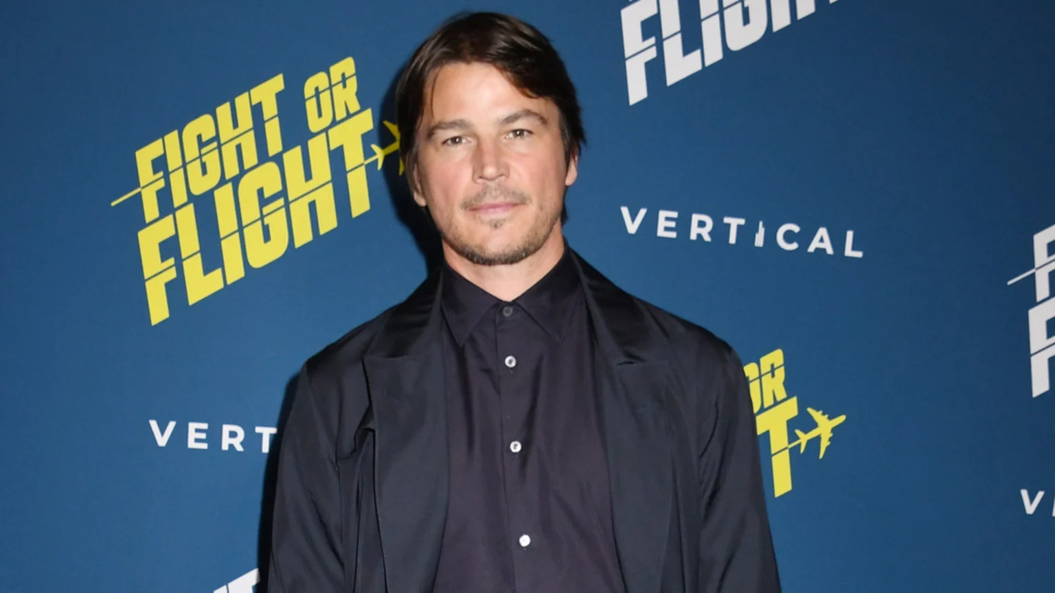 Josh Hartnett in a dark shirt and blazer poses against a blue background featuring "FIGHT OR FLIGHT" and "VERTICAL" logos.