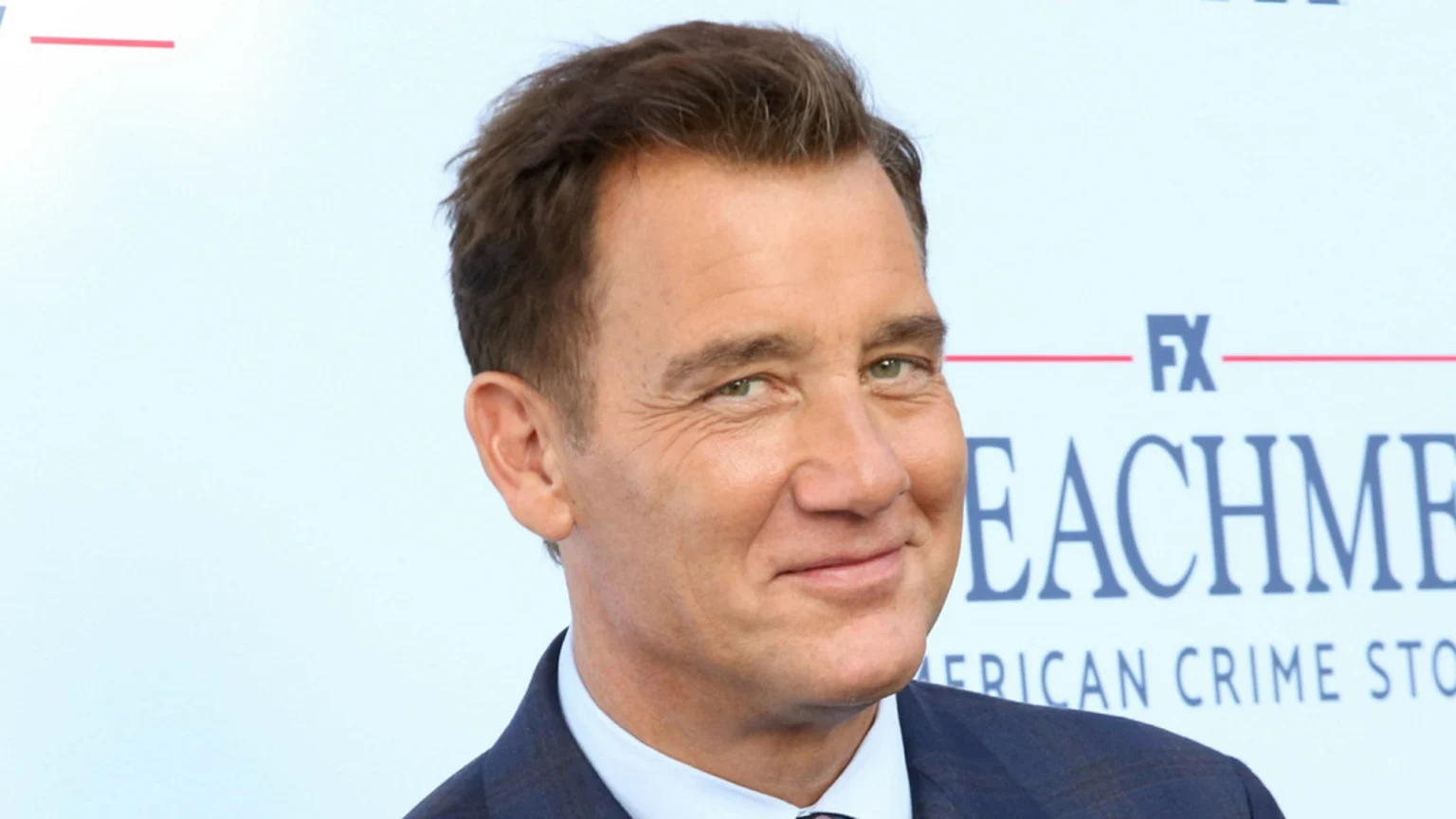 Actor Clive Owen, a man with brown hair and green eyes, wearing a dark suit, smiles faintly with "FX" and "IMPEACHMENT" visible in the white background.