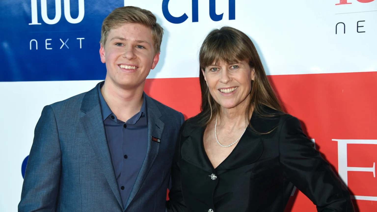 Robert Irwin and Terri Irwin smile side-by-side at an event, with Robert in a blue suit and Terri in a black jacket and pearl necklace.