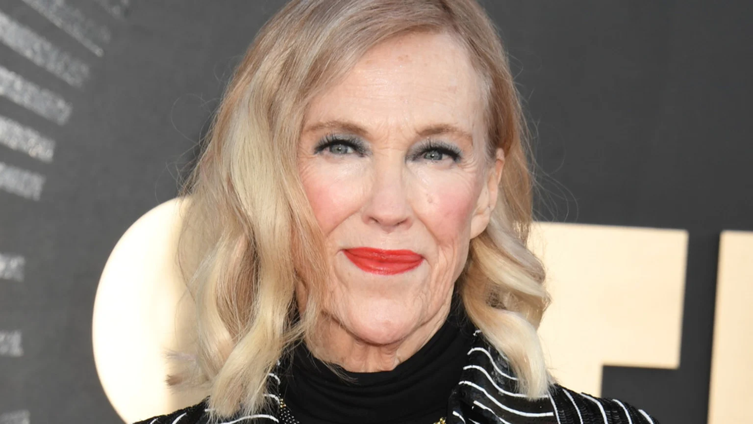 A blonde woman with wavy hair, blue eyeshadow, red lipstick, and visible wrinkles looks forward, wearing a black and white striped top.