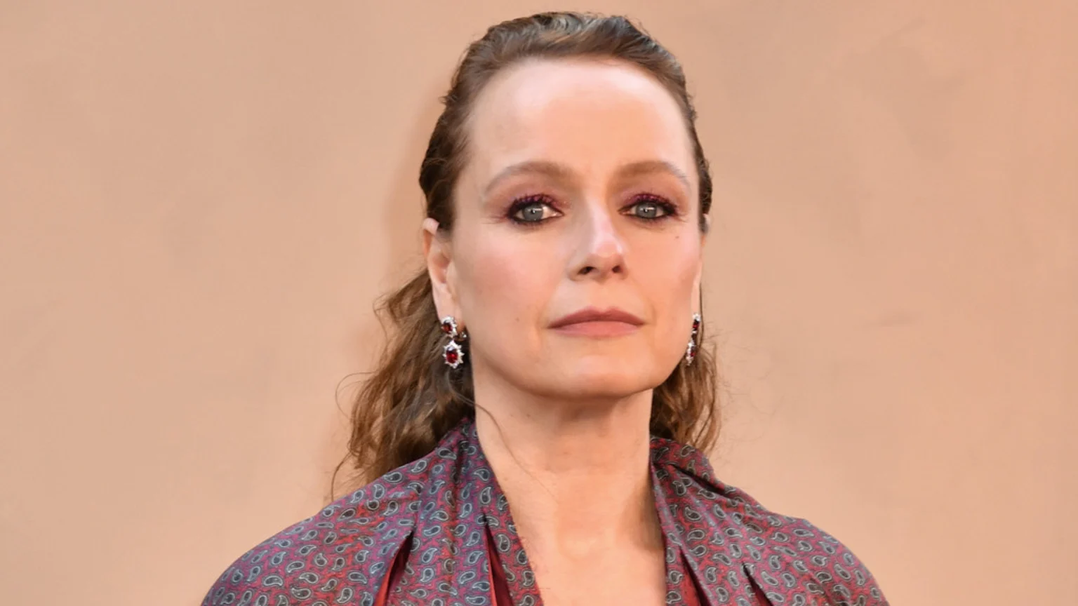 Actress Samantha Morton looks at the camera in a close-up, wearing bold red eyeshadow, red and silver drop earrings, and a dark red patterned top.