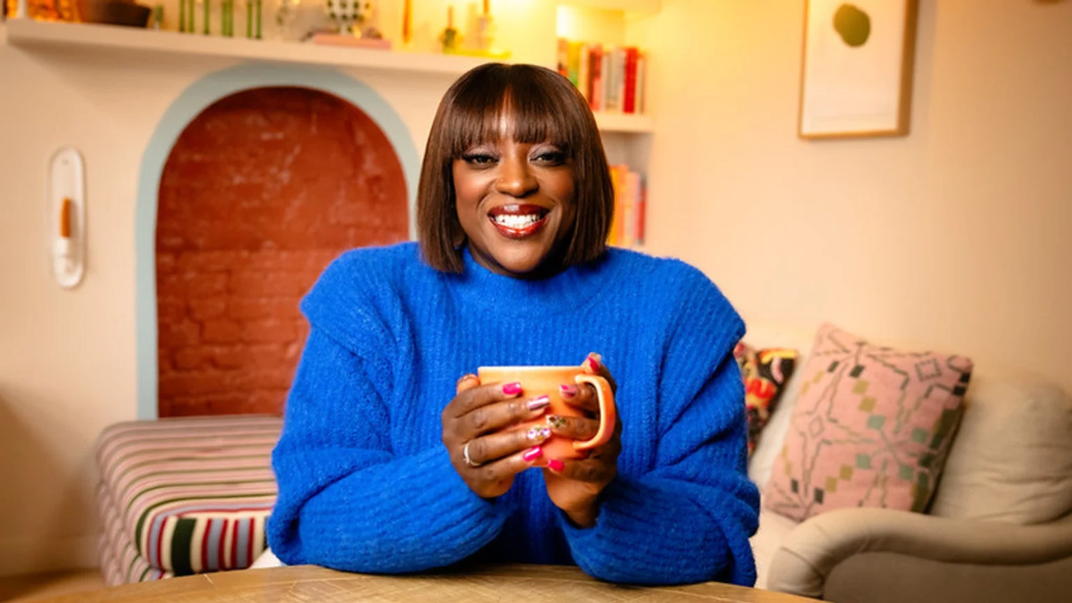 A smiling Black woman with a bob and bangs, wearing a blue sweater, holds an orange mug while looking directly at the viewer in a cozy, warmly lit room.