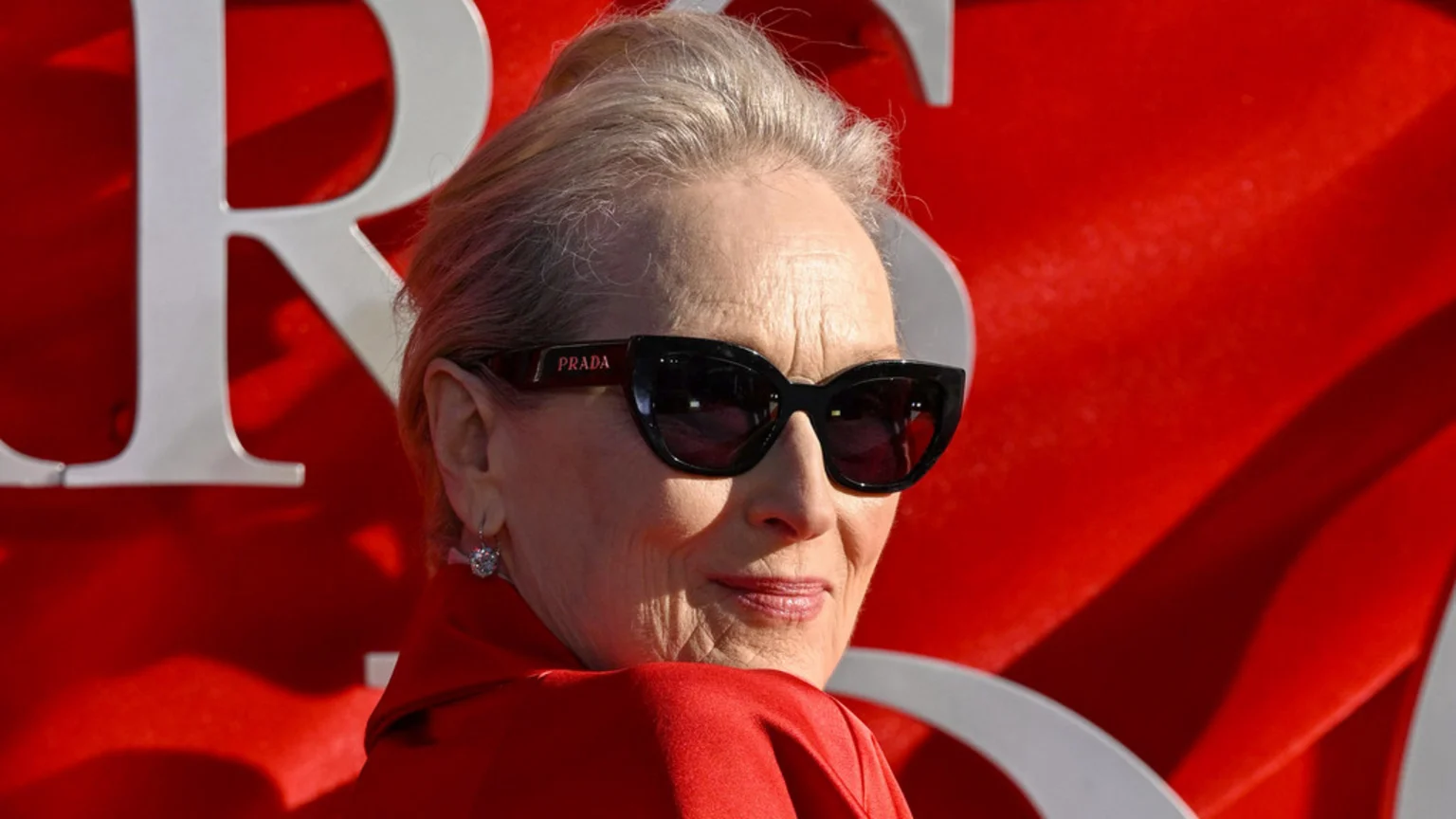 Meryl Streep, wearing black Prada sunglasses and a red outfit, smiles slightly while looking over her shoulder against a red background with white letters.