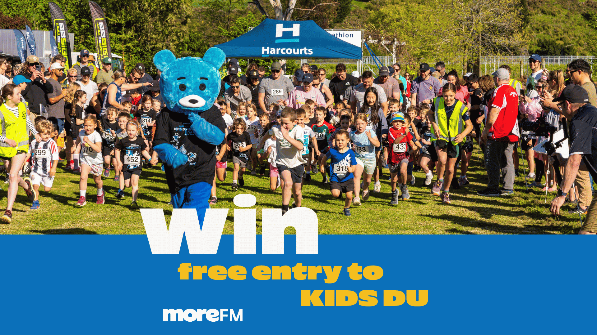 A blue mascot bear and many children run a race on a grassy field, with adults watching, a "Harcourts" tent, and "Win free entry to KIDS DU" text.
